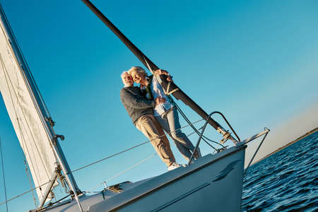 Full length of a romantic and happy senior couple standing on the side of sail boat or yacht deck floating in the calm blue sea, hugging and enjoying amazing sunsetの写真素材