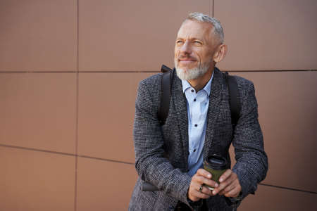 Modern middle aged businessman looking thoughtful while standing outdoors with electric scooter and a cup of coffeeの写真素材