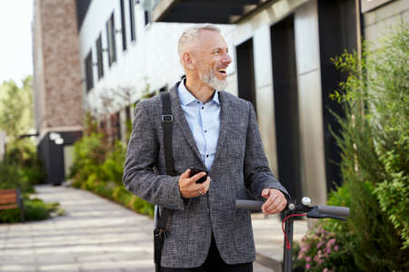 Joyful mature businessman holding smartphone, looking aside while walking with electric scooter outdoors on a daytimeの写真素材