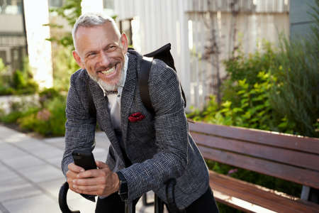 Happy middle aged businessman in stylish suit smiling while holding his smartphone, standing with bicycle outdoors in the daytimeの写真素材