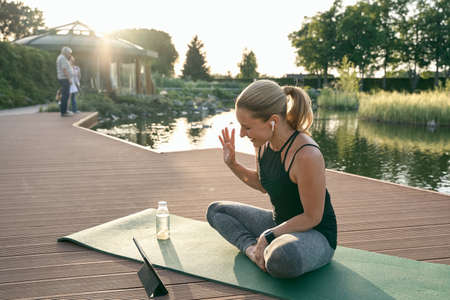 Online workout. Happy sporty woman waving while making a video call using digital tablet, doing yoga on a mat in nature on a sunny dayの写真素材