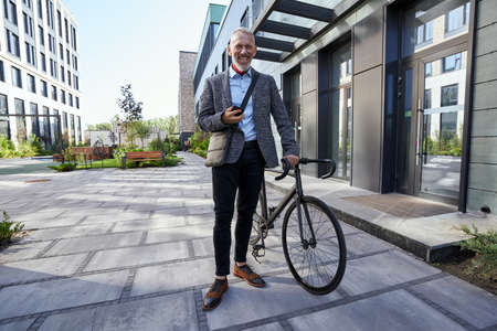 Full length shot of stylish mature businessman smiling while using his mobile phone, standing with bicycle near modern office building outdoorsの写真素材