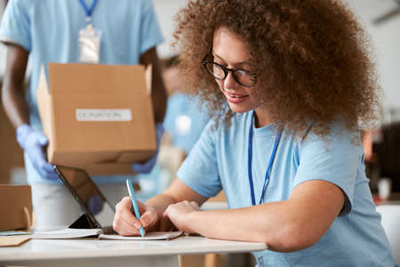 Young woman volunteer making notes and guy carrying cardboard box in the background while working on donation project indoorsの写真素材