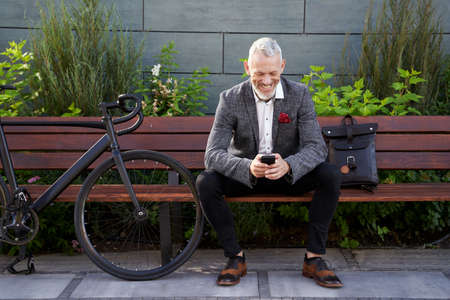 Resting. Successful middle aged business man in stylish suit texting, using mobile phone while sitting on the bench outdoors with a bicycle next to himの写真素材