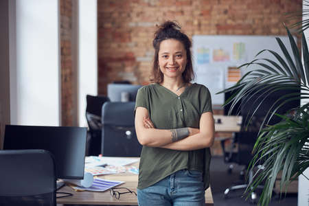Portrait of happy woman, interior designer or architect smiling at camera while standing with arms crossed in modern green office on a daytimeの写真素材