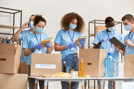 Young team of volunteers in protective masks and gloves sorting, packing food and water in cardboard...