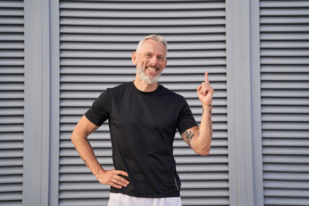Cheerful mature man smiling at camera, raising hand and pointing index finger up while posing outdoors over gray backgroundの写真素材