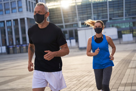 Sporty middle aged couple wearing protective masks running in urban environment, staying active during quarantineの写真素材