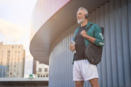 Happy sportive middle aged guy in sportswear and headphones looking away, standing outdoors, ready for tennis workoutの写真素材