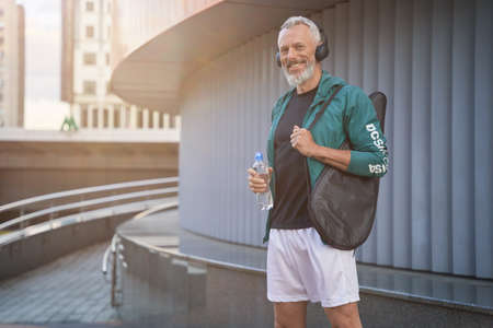 Happy sportive middle aged man in sportswear and headphones smiling at camera, standing outdoors, ready for tennis workoutの写真素材