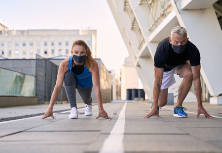 Active mature couple wearing protective masks on the start position ready for running together in urban environmentの写真素材