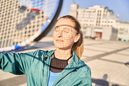 Portrait of attractive middle aged sportswoman holding tennis racket, standing with eyes closed, ready for morning workout outdoorsの写真素材