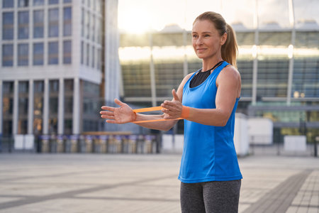 Beautiful sportive woman in sportswear, female trainer holding resistance band while standing outdoors, working out in the cityの写真素材