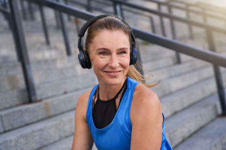 Portrait of active middle aged woman in sportswear and headphones smiling at camera, posing outdoors after morning workout in the cityの写真素材