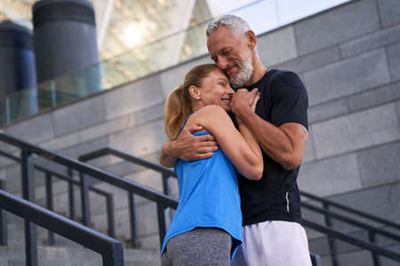 Affectionate middle aged couple, man and woman in sportswear embracing each other while standing together outdoorsの写真素材