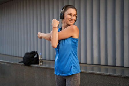 Sportive mature woman in sportswear and headphones smiling away while warming up, stretching her arms, standing outdoorsの写真素材