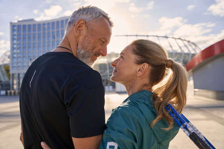 Lovely sportive middle aged couple smiling at each other while standing together outdoors, ready for morning workout on tennis courtの写真素材