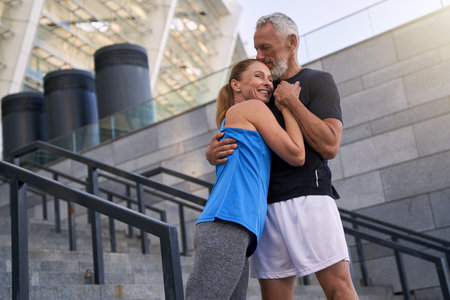 Lovely sportive middle aged couple, man and woman in sportswear hugging while standing together outdoorsの写真素材