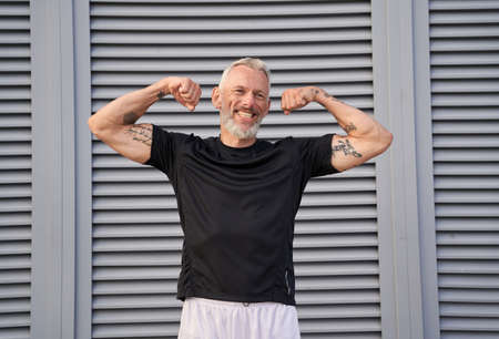 Smiling middle aged man raised his arms and shows his biceps while posing outdoors over gray backgroundの写真素材