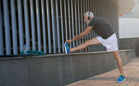 Full length shot of sportive middle aged man in sportswear and headphones warming up his body,...