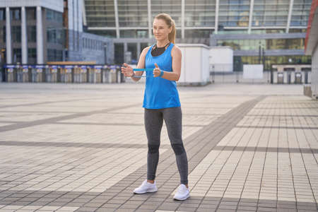 Full length shot of smiling sporty woman in sportswear, female trainer holding resistance band while standing outdoors, exercising in the cityの写真素材