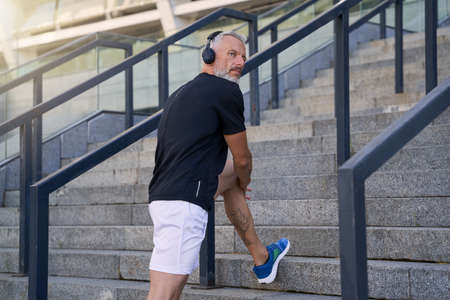 Fit middle aged man in sportswear and headphones listening to music or podcast, looking aside while exercising outdoorsの写真素材
