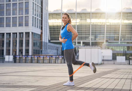 Full length shot of smiling sporty woman in sportswear using resistance band while standing outdoors, exercising in the cityの写真素材