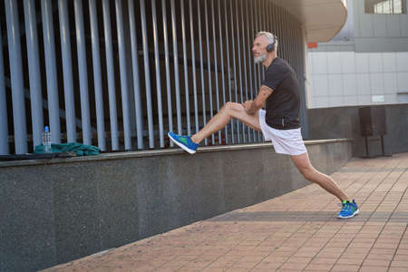 Full length shot of sportive middle aged man in sportswear and headphones looking away, warming up his body, getting ready for workout outdoorsの写真素材