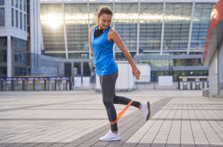 Full length shot of active sporty woman in sportswear warming up using resistance band while standing outdoors, exercising in the cityの写真素材