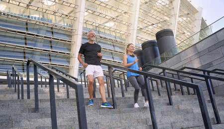 Cheerful active couple, man and woman in sportswear having workout together outdoors, jogging down the stairs in cityの写真素材