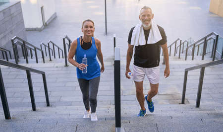 Joyful middle aged couple, man and woman in sportswear smiling at camera, walking up the stairs...