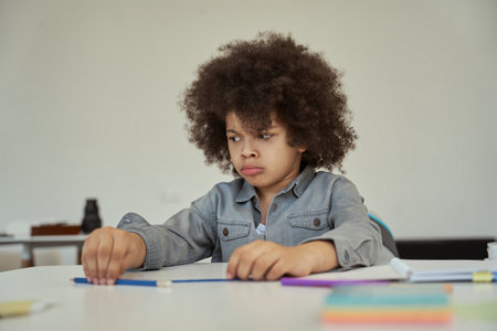 Disgruntled little schoolboy with afro hair looking sad while sitting at the table in elementary school classroomの写真素材