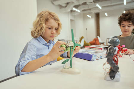 Create things. Caucasian boy examining technical toy while sitting at the table together with other kids, having engineering classの写真素材