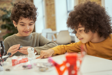 Kind little boy looking joyful while sitting at the table with his friend and examining details of a...