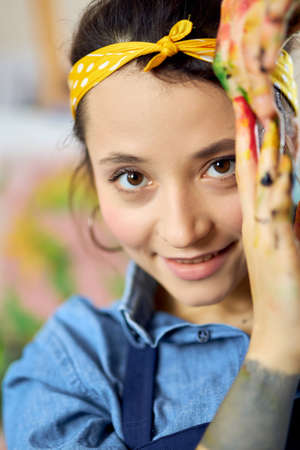 Close up portrait of happy young woman, female artist looking at camera, posing with her hands in paint while creating paintingの写真素材