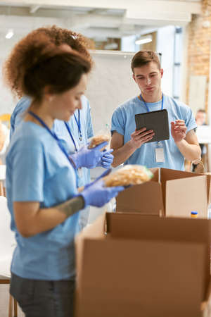 Young guy calculating using tablet pc. Volunteers in protective gloves sorting, packing food in cardboard boxes, working together on donation project indoorsの写真素材
