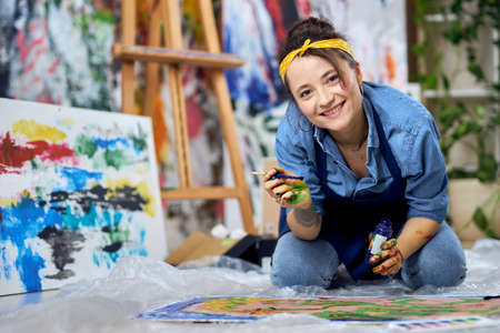 Happy young woman in apron smiling at camera, holding paintbrush while working on painting, sitting on the floor at home studio workshopの写真素材