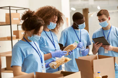 Young team of volunteers in protective masks and gloves sorting, packing food in cardboard boxes, working on donation project indoorsの写真素材