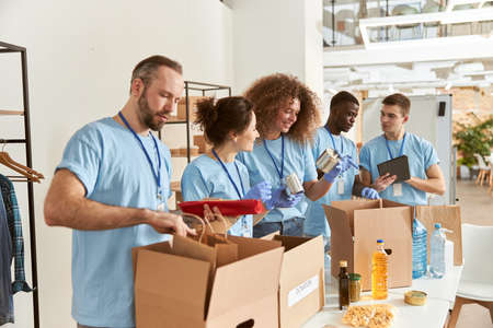 Team of diverse volunteers in protective gloves sorting, packing foodstuff in cardboard boxes, working together on donation project indoorsの写真素材