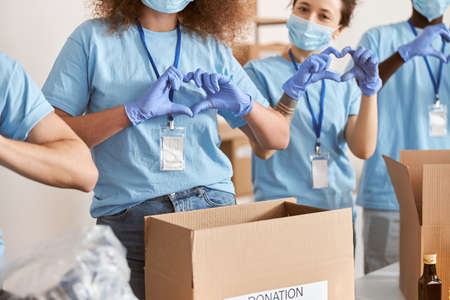 Cropped shot of diverse people wearing blue uniform, protective masks and gloves showing love heart sign while volunteering in community, sorting donated food itemsの写真素材