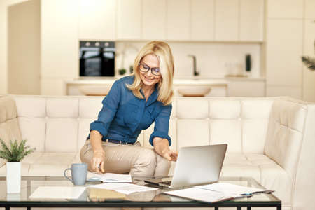 Joyful mature business woman in casual wear making notes, working from home while sitting on a couch in front of laptop in modern apartmentの写真素材