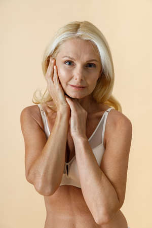 Portrait of natural mature woman with blonde hair in white underwear looking at camera, touching her face while posing isolated over beige backgroundの写真素材