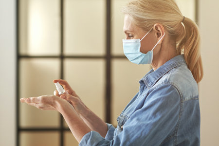 Side view of mature blonde woman wearing protective face mask applying antibacterial hand sanitizer gel pump dispenser while cleaning her handsの写真素材