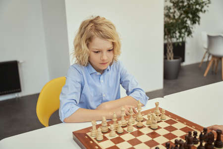 Intellectual game. Smart caucasian boy in blue shirt sitting in the classroom and playing chess on the chessboardの写真素材