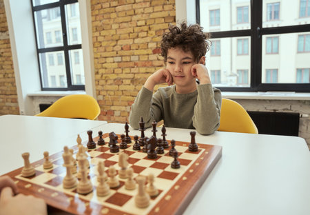 Observant little boy sitting in the classroom and playing chess on the chessboardの写真素材