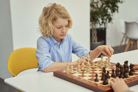 Looking for a good move. Curious little caucasian boy in blue shirt sitting in the classroom and playing chess on the chessboardの写真素材