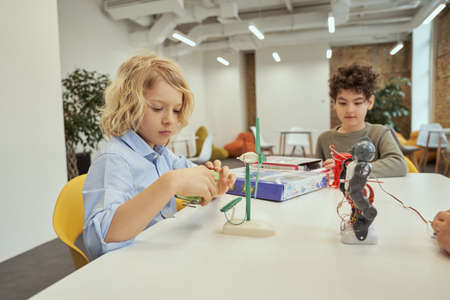 Curious caucasian boy examining technical toy full of details while sitting at the table together with other kids, having engineering classの写真素材