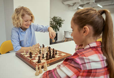 Playful little caucasian boy smiling while making his move, playing chess with friend, sitting together at the table in schoolの写真素材