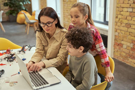 Attractive young female teacher in glasses showing scientific robotics video to children, using...