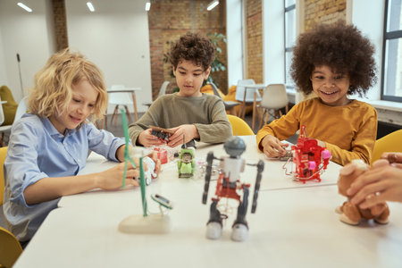 Innovations. Curious little diverse boys smiling while sitting at the table and checking robot toys, spending time together at engineering clubの写真素材